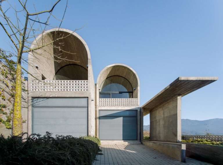 Low-angle view of a textured raw concrete wall and cantilevered roof canopy at Casa Cielo above wild grasses and green landscaping.