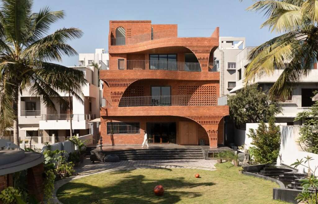 Top-down aerial drone view of the organic residence, featuring a circular green lawn, paved stone paths, palm trees, and the shifting geometry of the roof terraces.