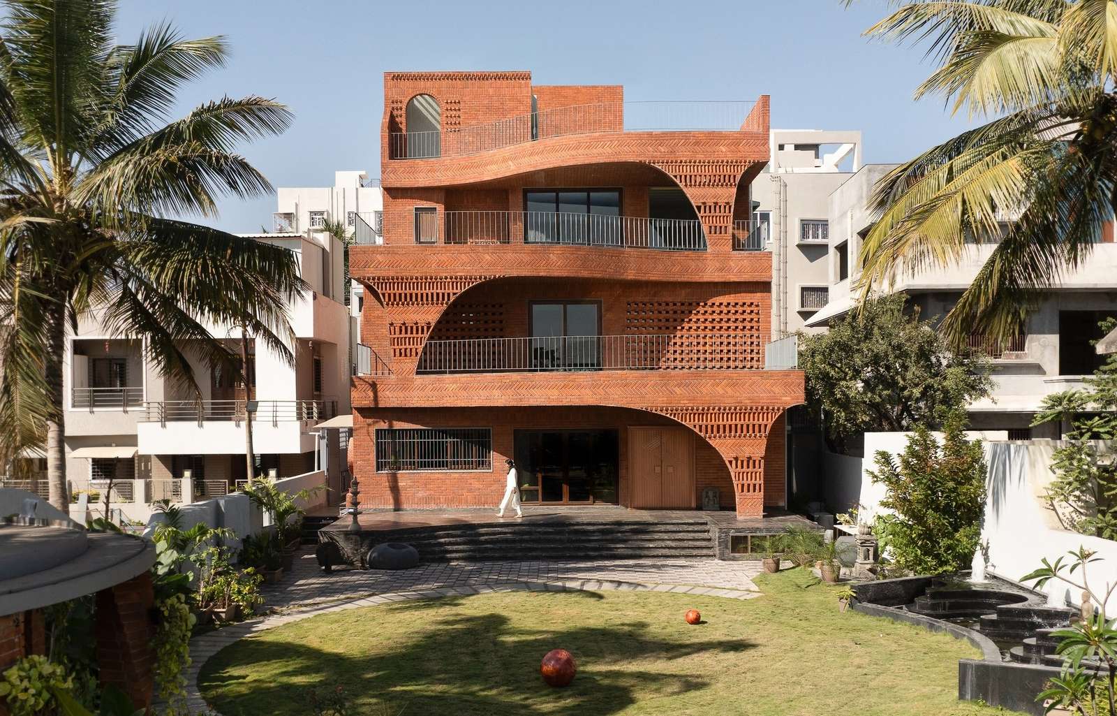 Top-down aerial drone view of the organic residence, featuring a circular green lawn, paved stone paths, palm trees, and the shifting geometry of the roof terraces.
