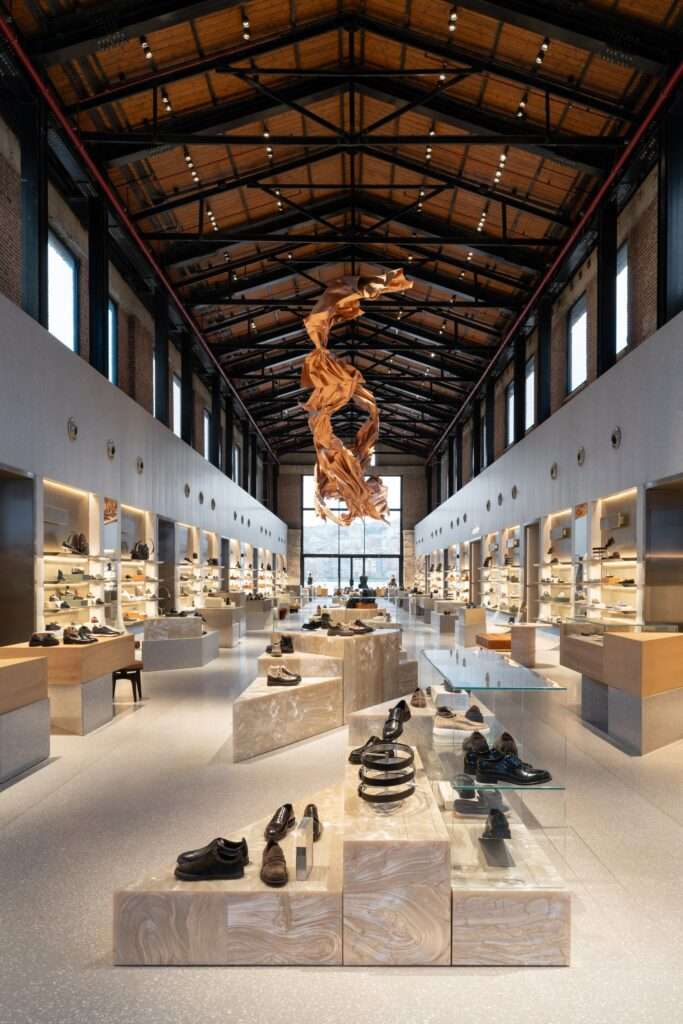 A long, double-height retail hall inside Beymen Tersane featuring an exposed wooden roof truss, geometric display islands, and a suspended fabric sculpture.