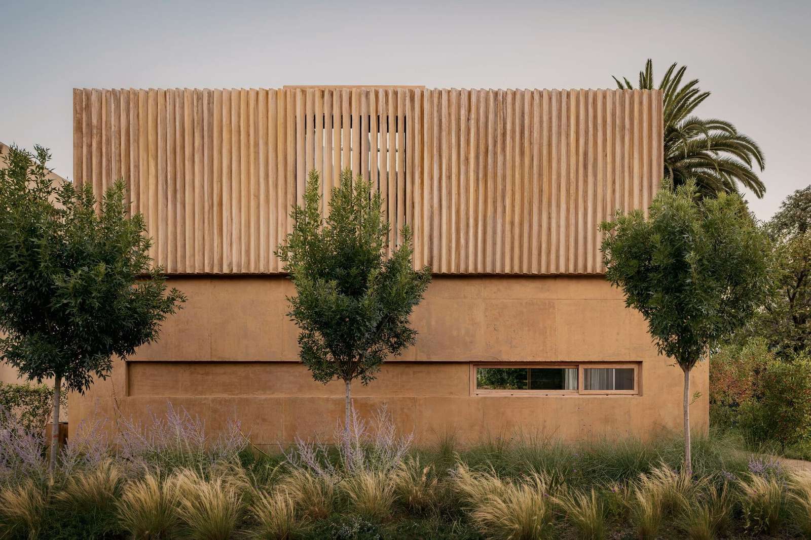 Exterior facade of JR House showing the textured concrete walls and vertical wooden slats blending with surrounding lush greenery.