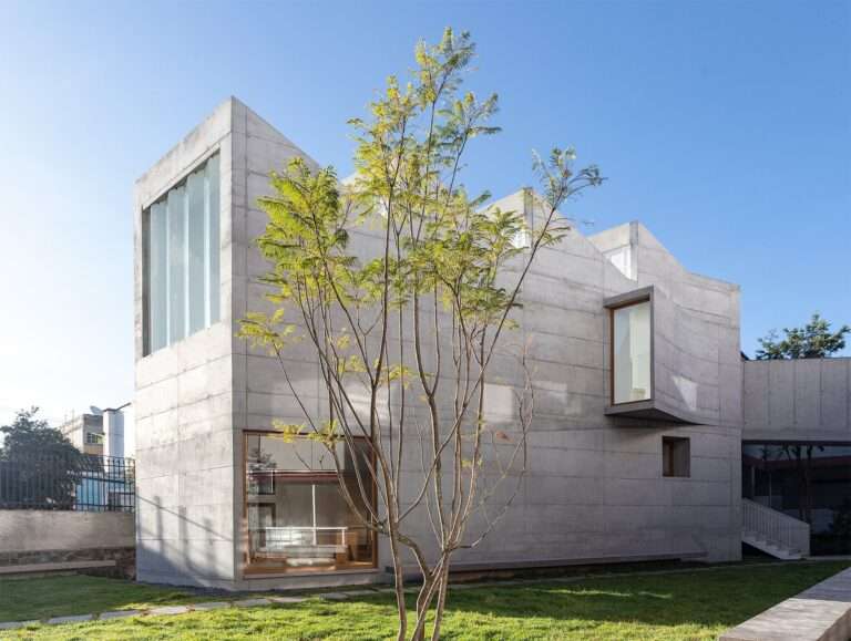 Exterior view of a modern board-formed concrete arts pavilion with large glass windows and an angular roofline, seen from a green lawn with a young tree.