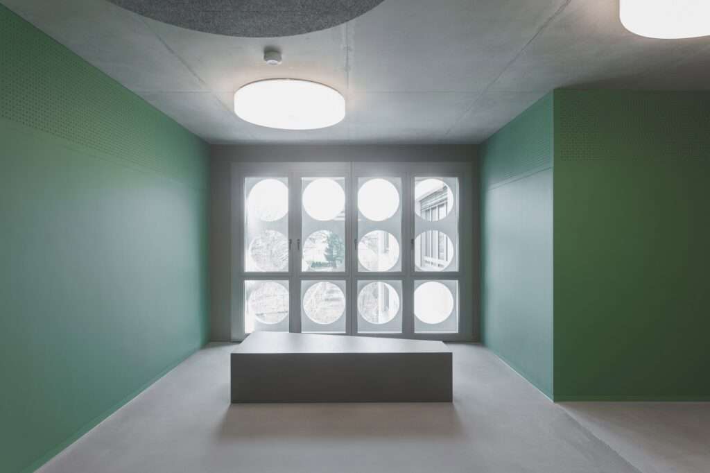Interior view of a school corridor with green walls, circular windows, and minimalist functional seating.
