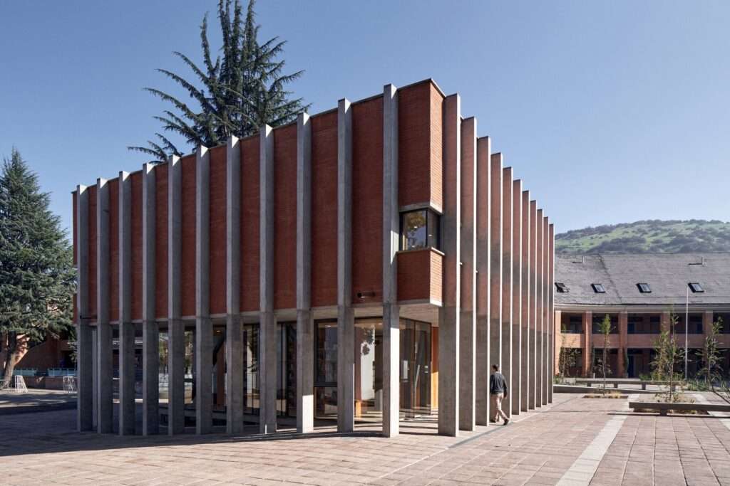 Modern two-story school library pavilion featuring a transparent glass ground floor beneath a solid monolithic upper brick volume enclosed by a regular grid of vertical concrete columns.