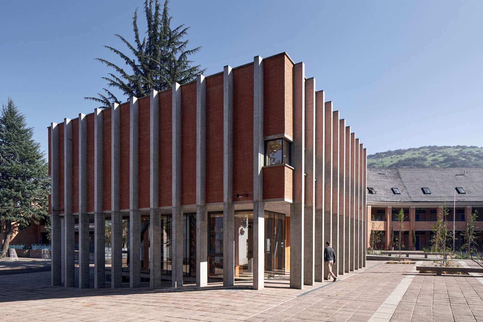 Modern two-story school library pavilion featuring a transparent glass ground floor beneath a solid monolithic upper brick volume enclosed by a regular grid of vertical concrete columns.