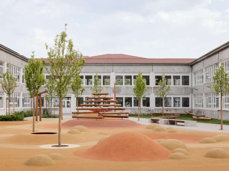 A colorful, textured children's playground area with mounds, climbing structures, and young trees in a school courtyard in Trnava.