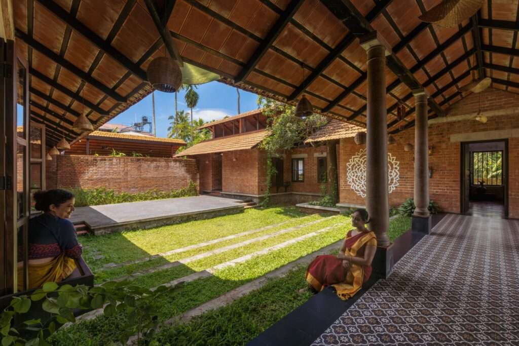 Interior view of a covered verandah with patterned floor tiles, wooden pillars, and women sitting, looking out onto an open grassy courtyard stage.