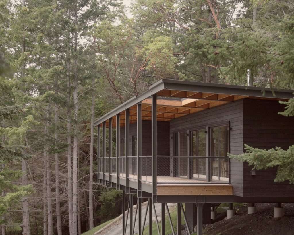 Side view of Trestle Cabin showing a dark wood modular volume suspended on a steel framework over a forested slope, with a deep wooden balcony roof.