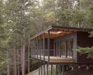 Side view of Trestle Cabin showing a dark wood modular volume suspended on a steel framework over a forested slope, with a deep wooden balcony roof.