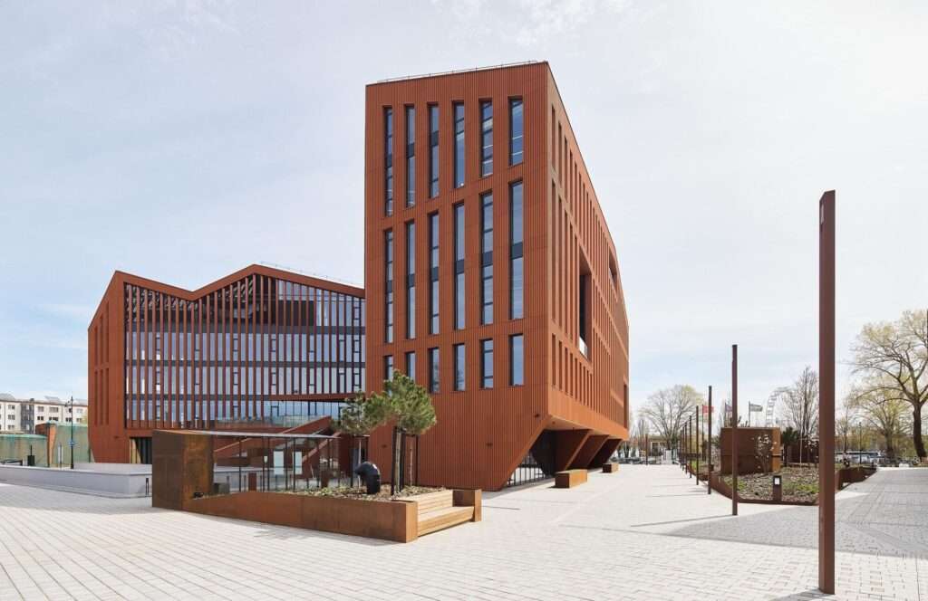 Angular perspective view of the triangular-shaped HANZA building showing a sharp corner cantilever, vertical terracotta louvers, and a wide-paved pedestrian walkway with integrated planters.