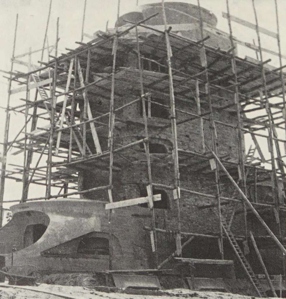Historical black and white photograph of the Einstein Tower under construction, completely covered in wooden scaffolding revealing its raw brickwork structure.