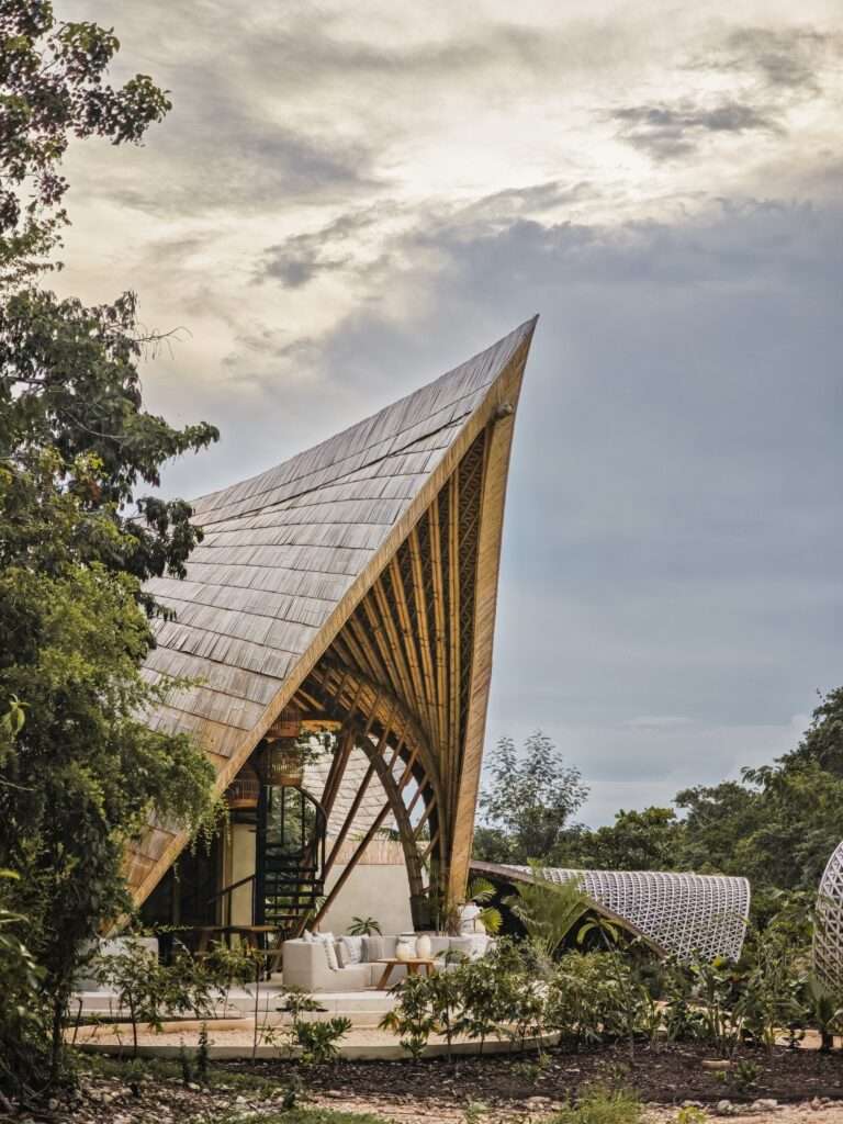 Ground-level exterior view of CORA Villa with a pointed bamboo roof structure towering over an outdoor seating area amid tropical vegetation under a cloudy sky.