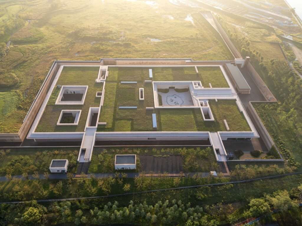 Aerial bird's-eye view of a modern sunken museum integrated into a green landscape with recessed open courtyards and grass-covered roofs at sunrise.