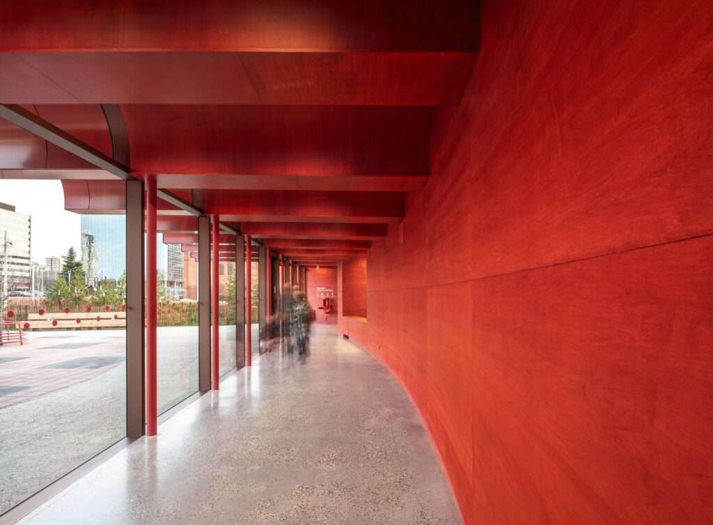 View from inside the O-day'min Park Pavilion showing the long corridor with vibrant red walls, polished concrete floors, and transparent glass facades.