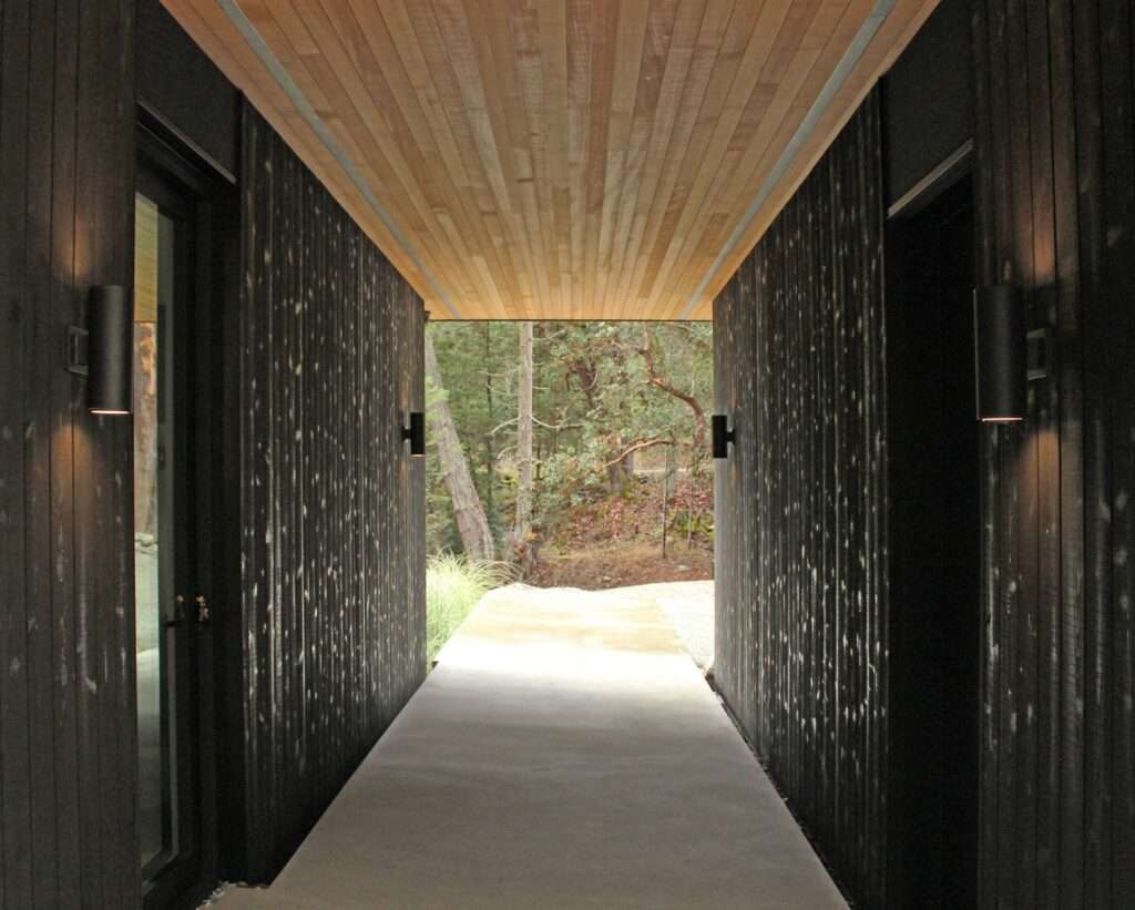 Symmetrical open-air concrete pathway hallway with dark textured wood walls under a warm wood plank ceiling looking toward a forest.