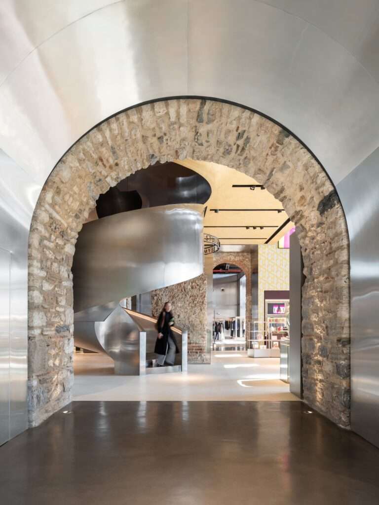 View through an ancient stone archway lined with a polished stainless steel frame, revealing a modern metallic spiral staircase inside.