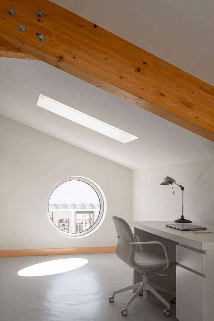 Minimalist white attic office with a sloped ceiling, an exposed wooden structural beam, a white desk with a laptop, a task lamp, and a circular porthole window reflecting an oval-shaped sunbeam on the floor.