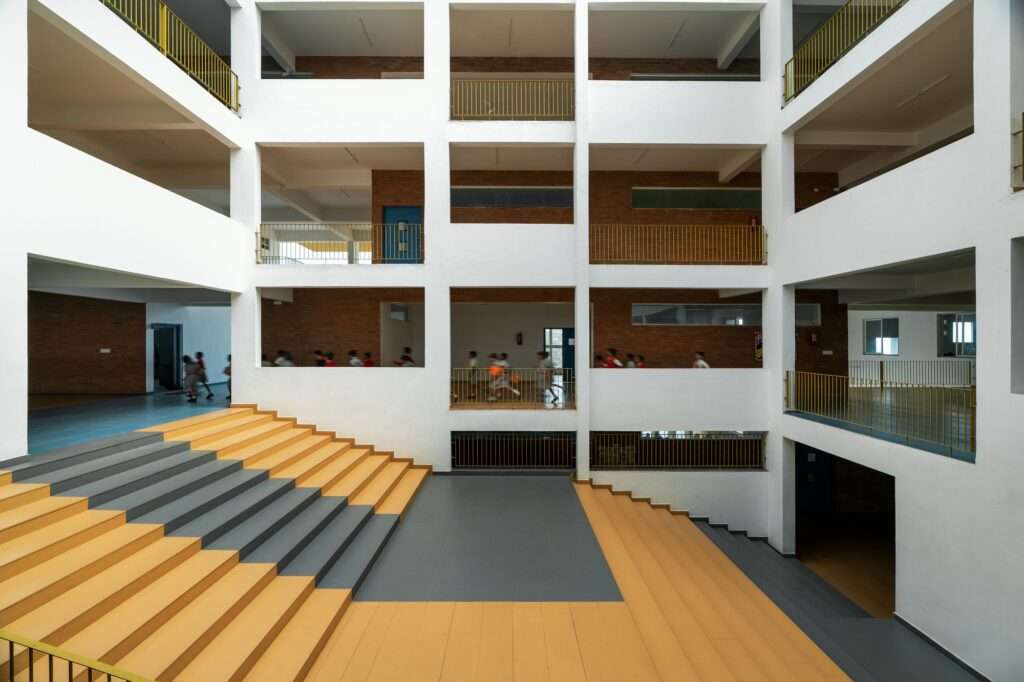 Interior courtyard amphitheater steps painted in yellow and grey within a school building featuring brick walls, concrete columns, and open corridors filled with moving students.