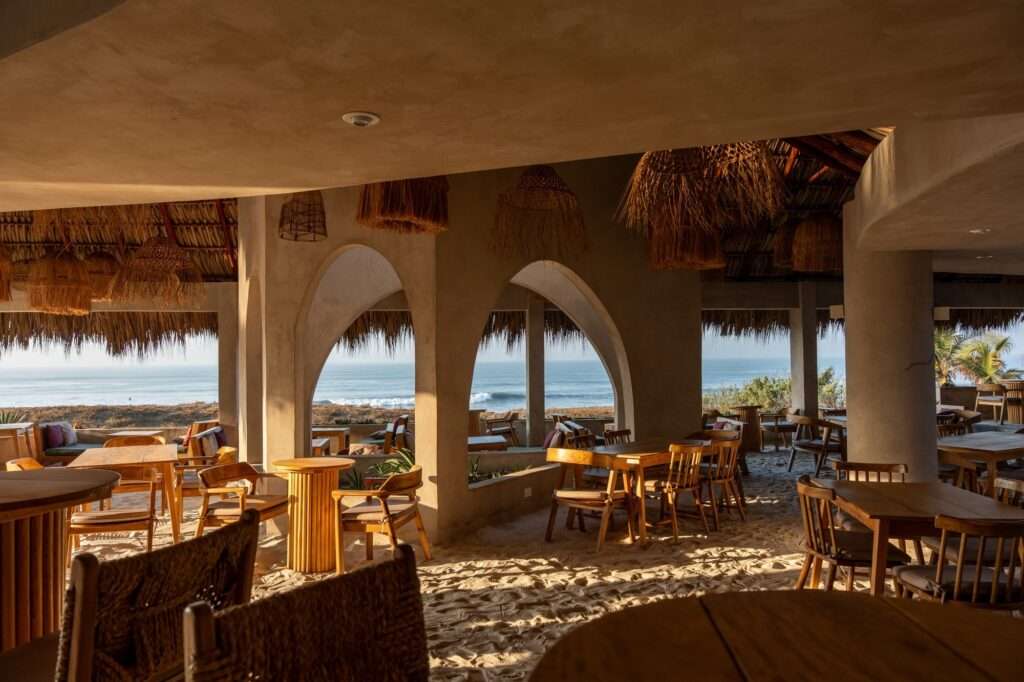 Interior view of a beach club restaurant featuring sand flooring, minimalist concrete arches framing an ocean view, and rustic wooden dining tables and woven chairs.