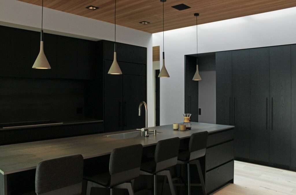 Minimalist dark gray kitchen island with bar stools under minimalist pendant lights with soft overhead skylight illumination.