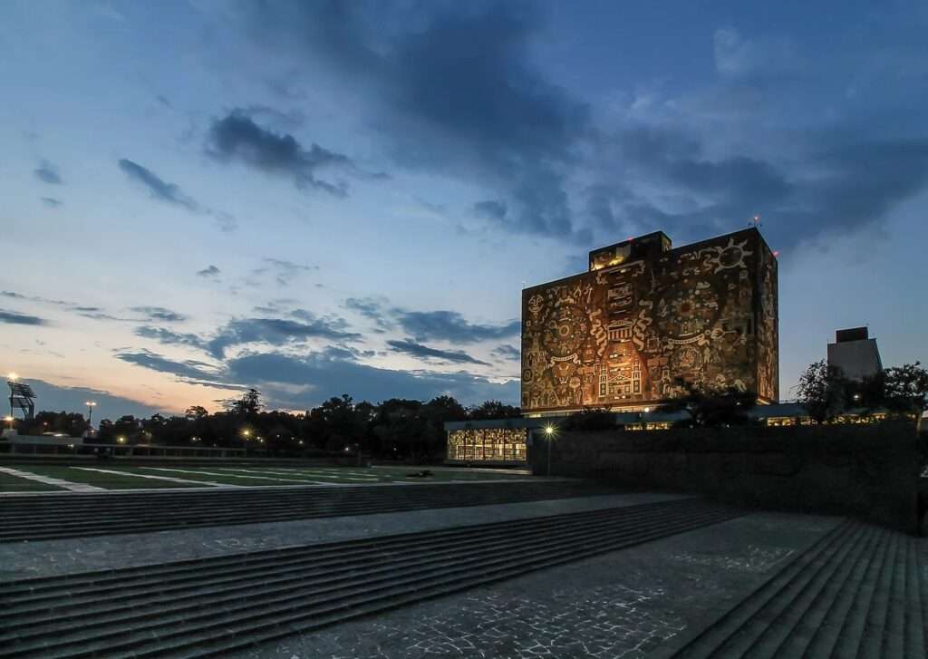UNAM Central Library building by Juan O'Gorman featuring the colorful historicist stone mosaic mural on the massive windowless archive tower rising above a transparent glass ground floor pavilion under a partly cloudy sky.