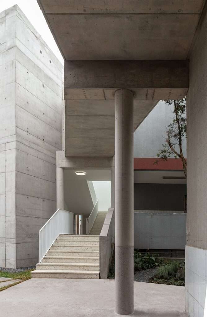 Exposed concrete structural connection showing an outdoor staircase, a connecting bridge, and a circular supporting column between two campus buildings.