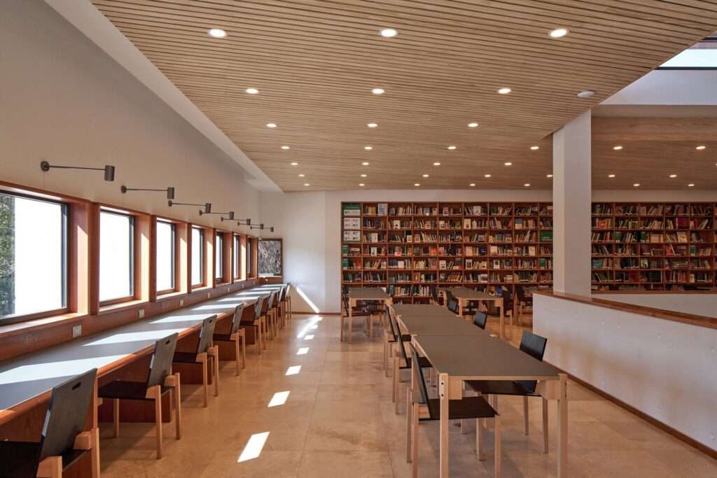Library interior reading room showing rows of study tables, a long built-in wooden perimeter desk beneath windows, warm timber slatted ceilings, and full-height bookshelves.