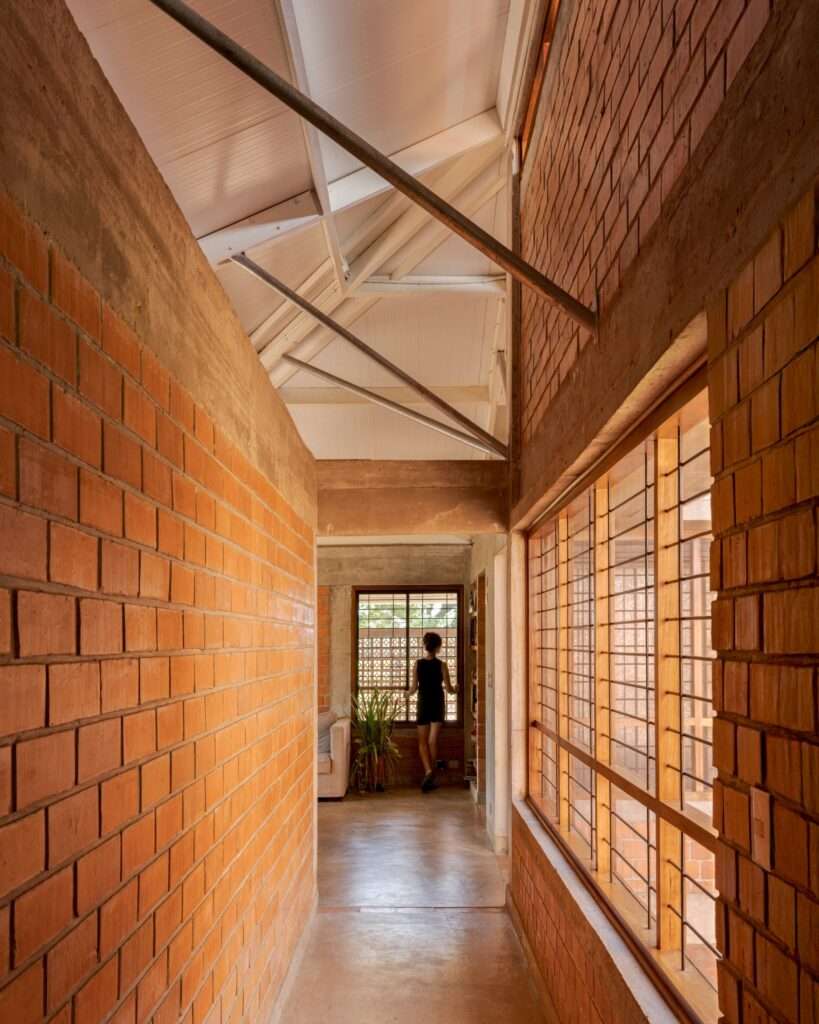 A bright interior corridor with red brick walls, concrete columns, and timber window frames looking toward a resident standing at the end.