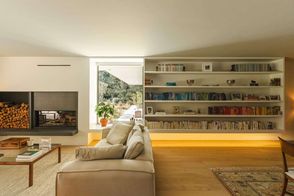 Modern open-plan living room interior featuring a large built-in bookshelf, minimalist sofa, and integrated fireplace facing an outdoor garden view.