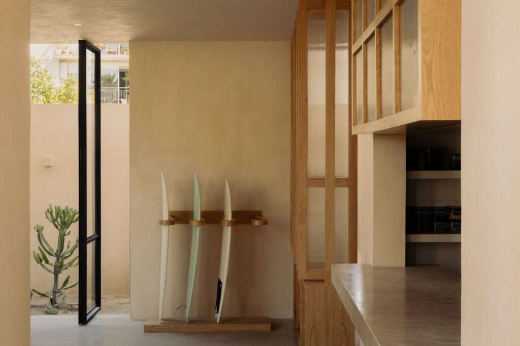 Interior corridor of Estero Residences featuring custom wooden cabinetry, a counter, a surfboard rack with three boards, and an open glass door looking out to a cactus garden.