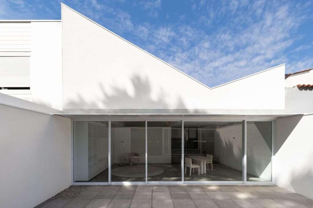 Minimalist white residential facade with a dynamic butterfly roof meeting at a central point above large sliding glass doors overlooking a concrete paved patio.