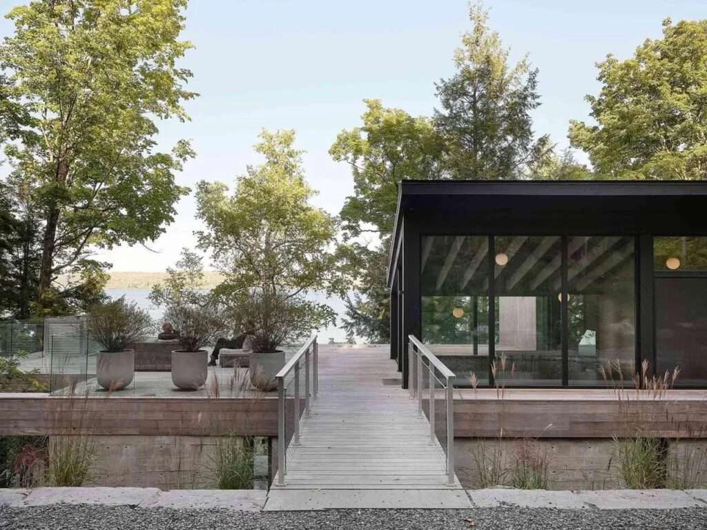 Modern residential building with glass walls and exposed wooden rafters, connected to an outdoor deck via a wooden pedestrian bridge surrounded by a dense forest near Lake Memphremagog.