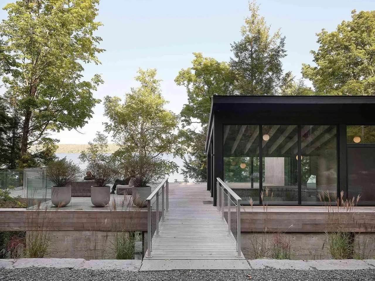 Modern residential building with glass walls and exposed wooden rafters, connected to an outdoor deck via a wooden pedestrian bridge surrounded by a dense forest near Lake Memphremagog.
