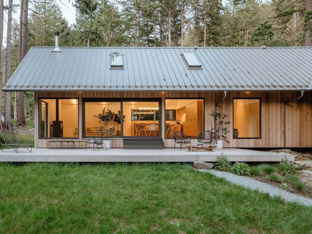 Daytime exterior view of the Floating House on Bowen Island, highlighting the natural cedar wood cladding, steep pitched metal roof with standing seams, large glass sliding doors, and an open wooden deck facing a lush green forest lawn.