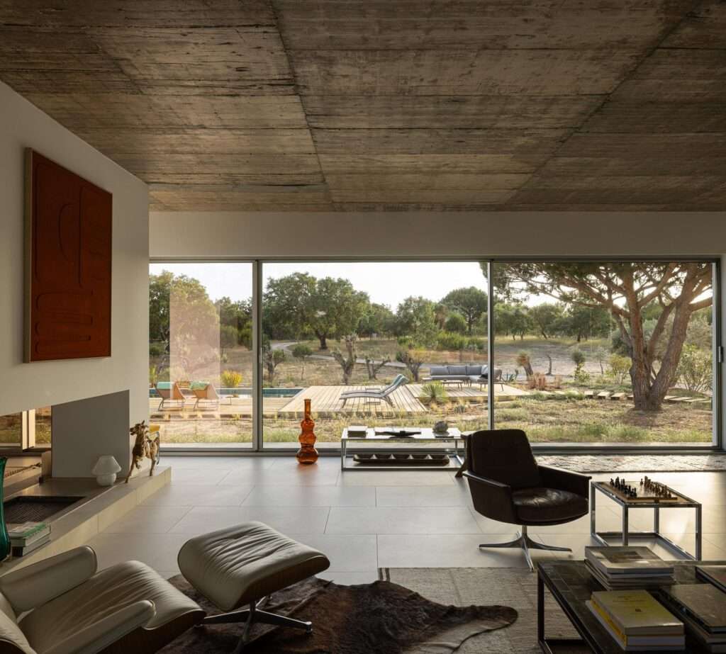 Spacious minimalist living room inside Melides House featuring a raw concrete ceiling, a white central fireplace with orange artwork, Vitra Eames lounge chair, and floor-to-ceiling glass doors overlooking the landscape.