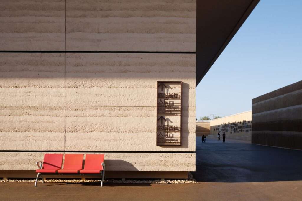 Exterior close-up of a modern building facade with texturized rammed-earth style finishes, directional signage, and a red outdoor bench.