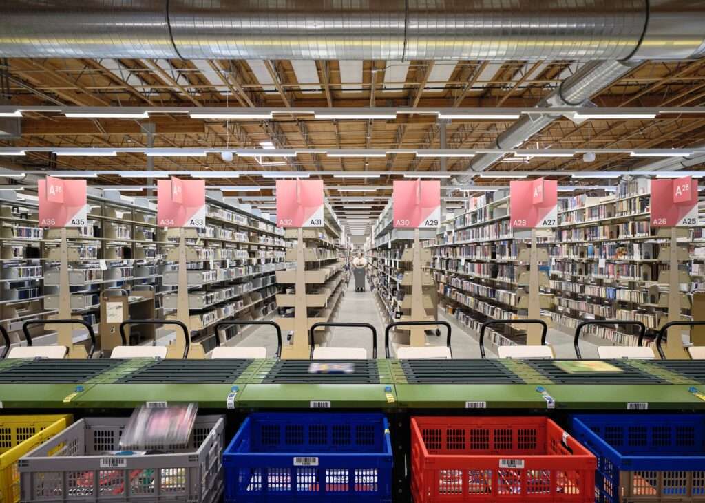 Interior view of the library operations facility showing automated conveyor belts sorting books into colorful plastic bins with high industrial book storage stacks under exposed wooden ceilings and ductwork.