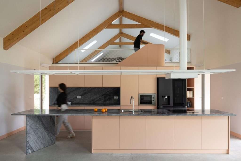 Contemporary kitchen with a minimalist dusty-pink cabinet wall, dark marble countertops and island, and a geometric staircase leading up to an open mezzanine attic.