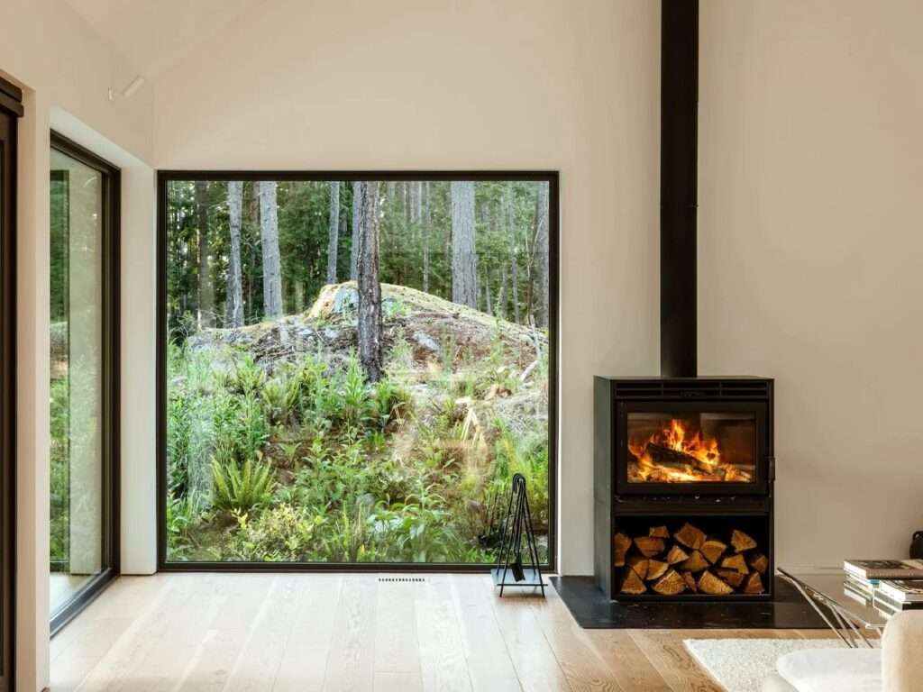 Interior view of a minimalist living room featuring light Douglas fir wooden flooring, a modern black steel wood-burning fireplace with stacked firewood underneath, and a massive floor-to-ceiling square window showcasing a rocky, moss-covered forest slope.