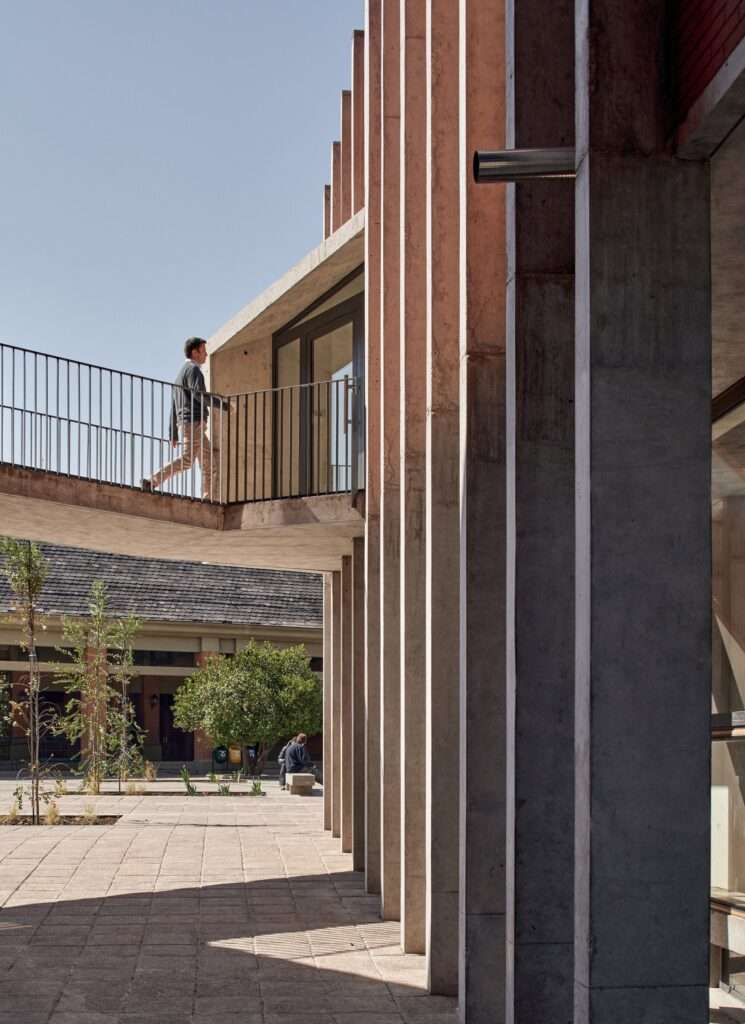 A pedestrian walking across a concrete elevated bridge that links the existing campus building to the upper level of the new library pavilion behind a screen of concrete fins.
