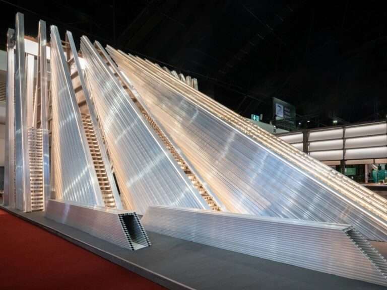 Exterior view of the UNFOLD Pavilion showcasing stacked layers of silver modular aluminum profiles forming a dynamic, sloped architectural structure under exhibition lighting.