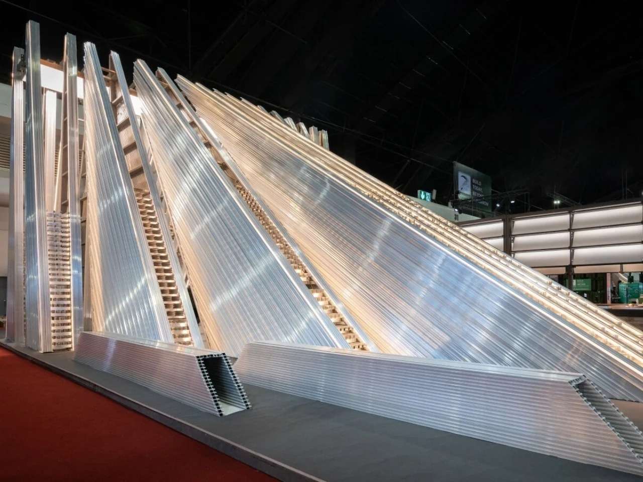 Exterior view of the UNFOLD Pavilion showcasing stacked layers of silver modular aluminum profiles forming a dynamic, sloped architectural structure under exhibition lighting.