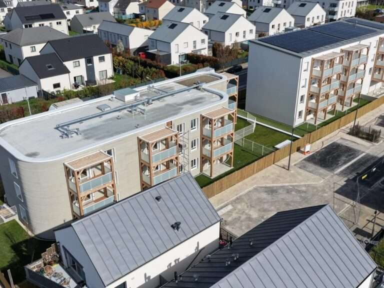 Aerial view of the ViliaSprint2 3D-printed residential building featuring curved concrete walls and wooden balconies next to a traditional building in Bezannes, France.