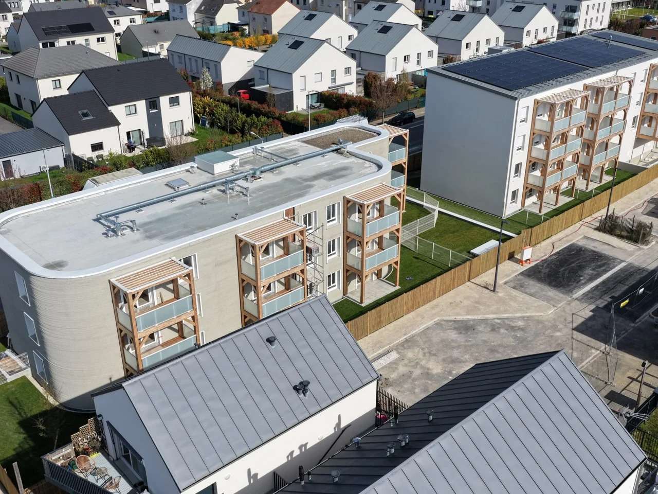 Aerial view of the ViliaSprint2 3D-printed residential building featuring curved concrete walls and wooden balconies next to a traditional building in Bezannes, France.