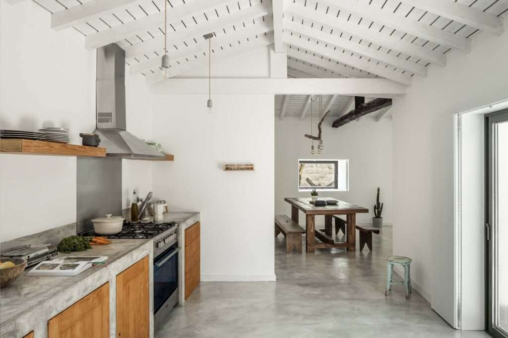 Contemporary kitchen featuring raw concrete countertops, warm wooden cabinetry, and stainless steel appliances beneath a white gabled ceiling, looking through to a rustic dining area.