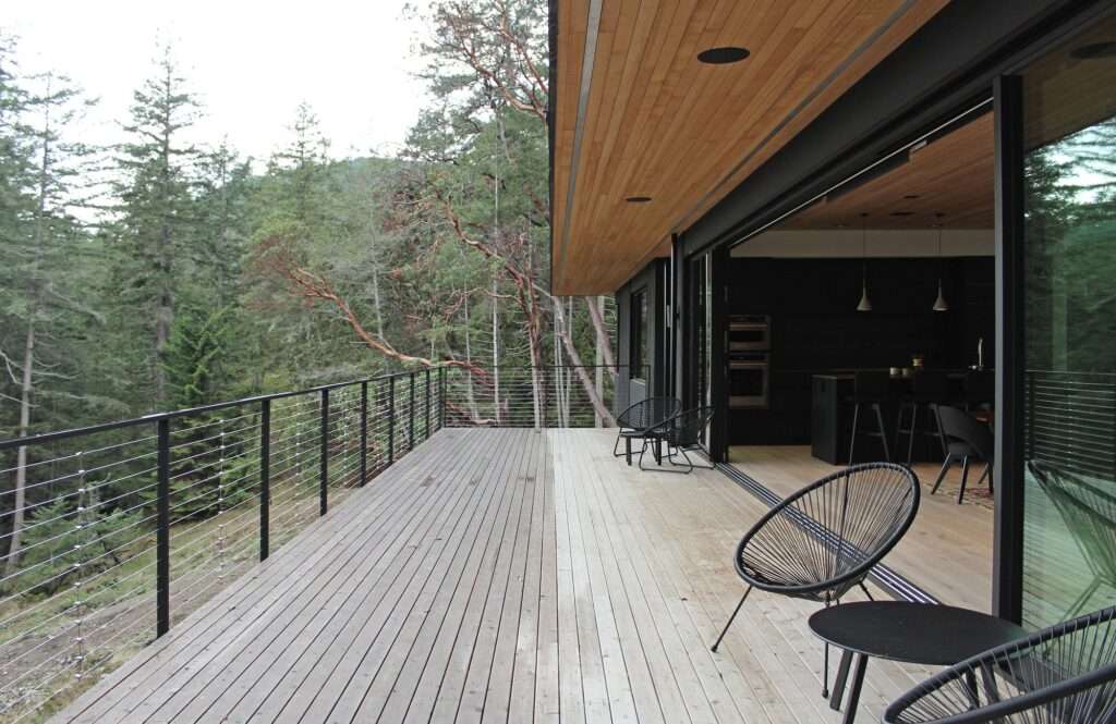 Large outdoor wooden deck balcony of Hilltop House with contemporary wire railings next to sliding glass doors facing a dense forest.