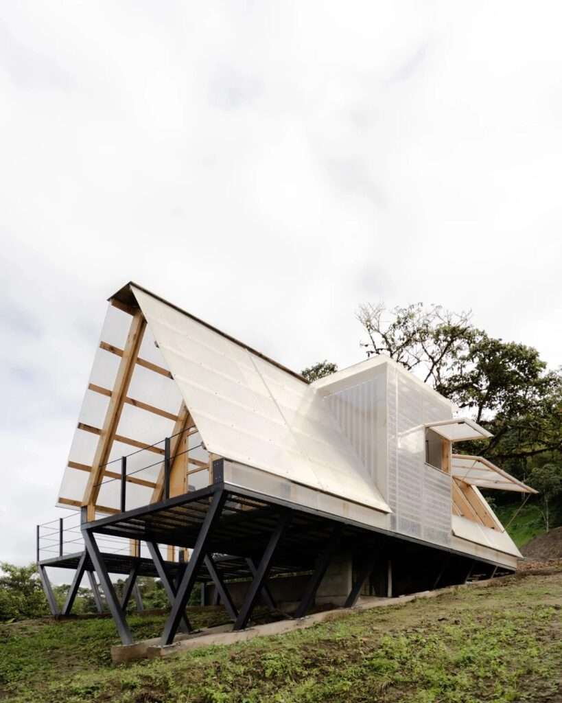 A daytime exterior view of Casa 6-3 cabin showing its distinctive A-frame roof and semi-transparent polycarbonate facade surrounded by dense green jungle trees.