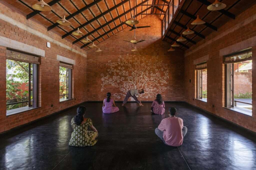 People practicing yoga in a spacious studio hall with exposed brick walls, polished dark flooring, and a large tree-of-life wall mural.