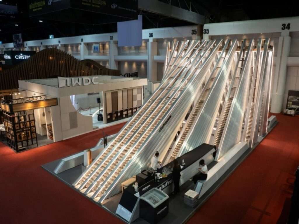 Elevated high-angle view of the UNFOLD Pavilion inside an exhibition hall, showing its linear, elongated wedge shape and open lounge area with visitors.