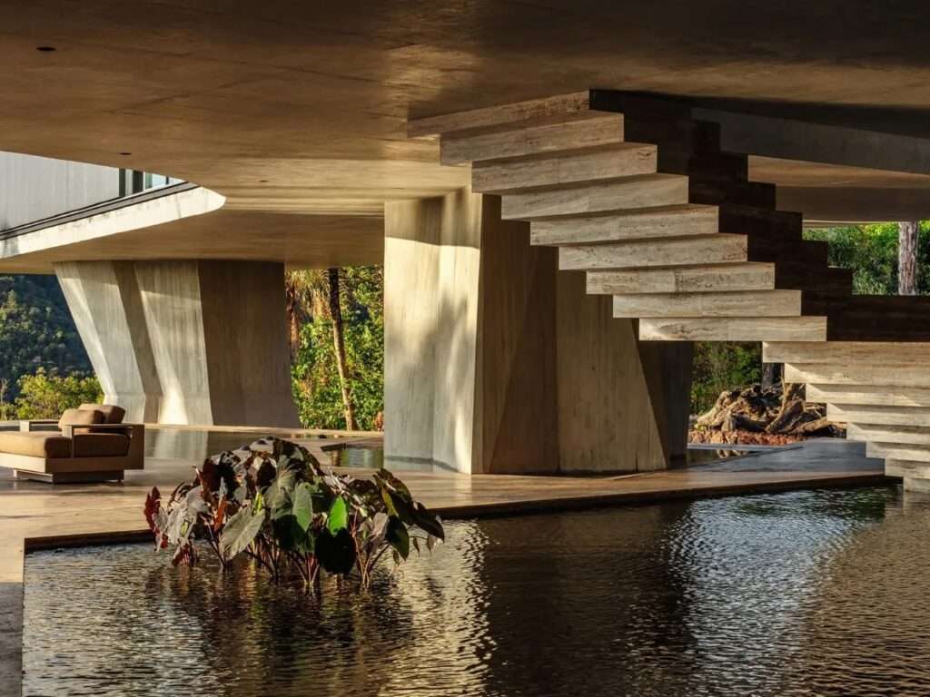 Modern cantilevered travertine stone staircase rising above a dark reflecting pool underneath a concrete building canopy.